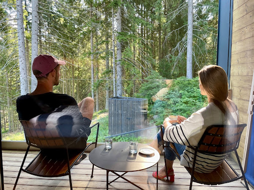 A man and a woman sit on a balcony, backs to the camera, staring into a forest.