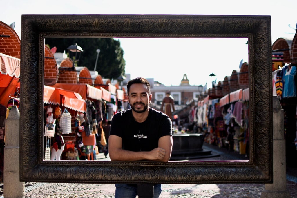 a young man poses for a photo behind a large picture frame, with a picturesque colorful market street on the background.