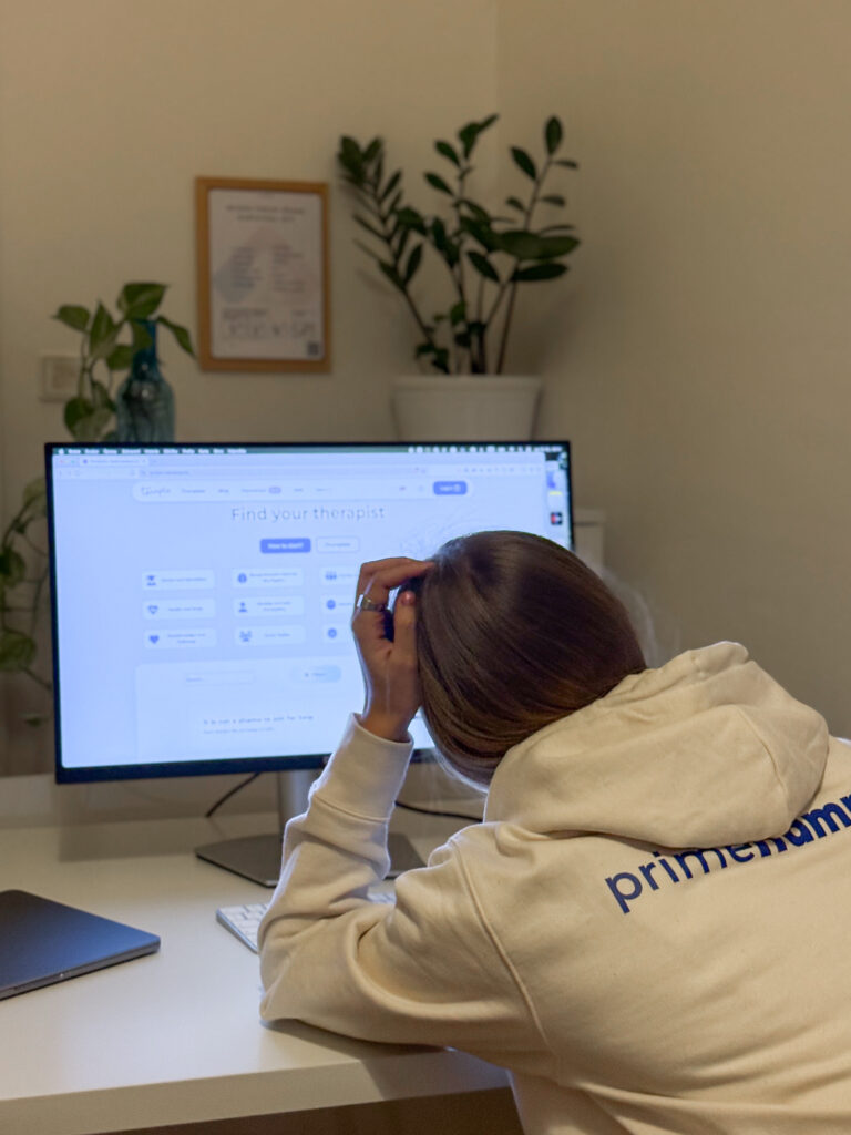 A blond woman in a white primehammer sweatshirt is seen from behind in front of a computer screen where she is booking a Terapio session. In the background there is a white office setting with two plants.