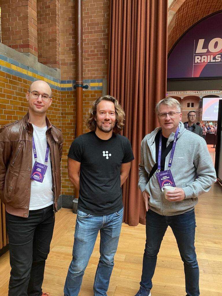 Three men pose for a picture at a Ruby on Rails conference: a primehammer engineer (a tall white man with a shaved head and glasses, wearing a brown leather jacket over a white T-shirt and black pants); DHH (a white man with shoulder-length dark-blond hair wearing a black T-shirt and jeans); and a slightly older-looking white man with short white hair, wearing a grey sweatshirt and dark jeans).