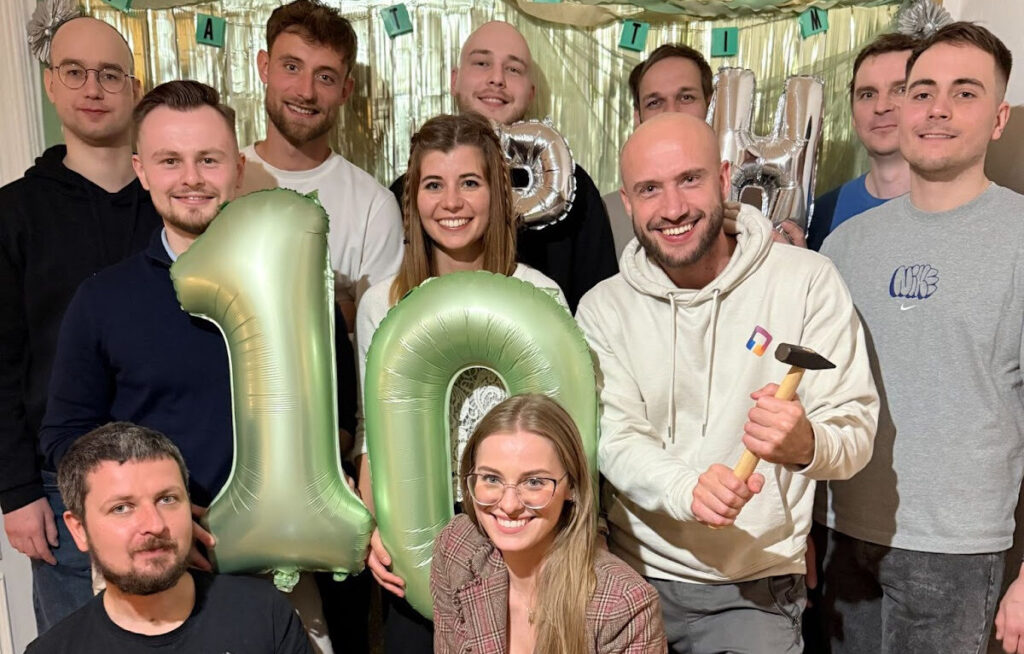 Eleven primehammer people pose for a group photo holding two green ballons (one shaped like a number 1, and the other shaped like the number 0, to make up the number 10).