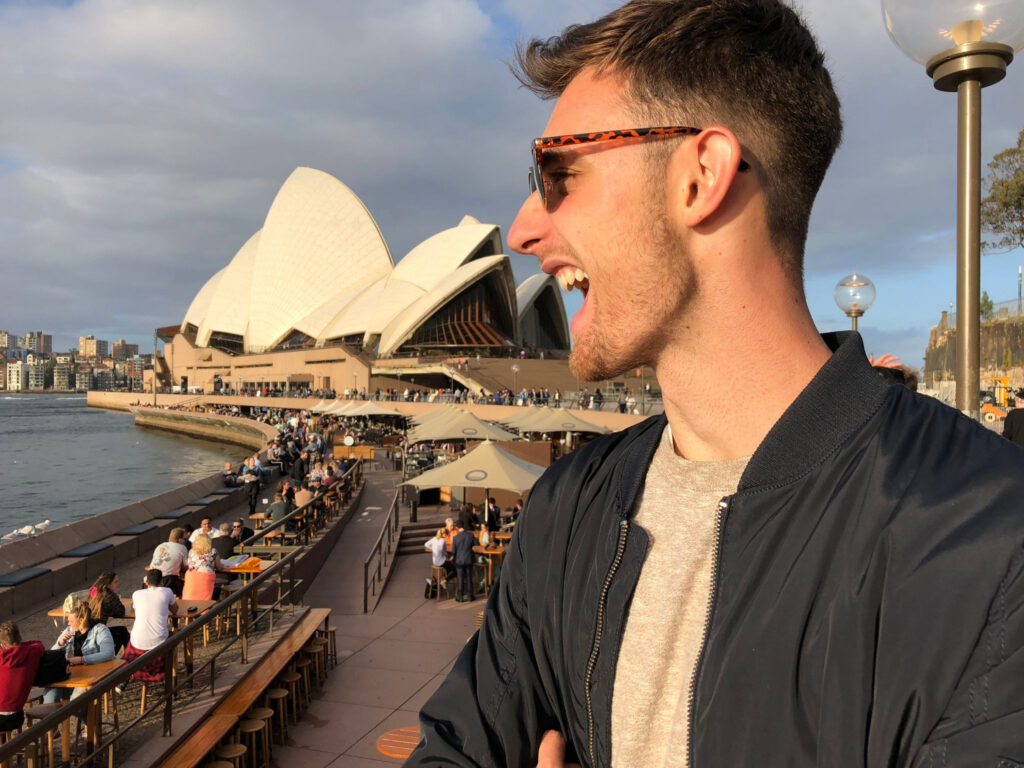 Photo of Daniel, a white man with brown hair, wearing sunglasses and his mouth open, pretending to eat the Sydney Opera House in the background.