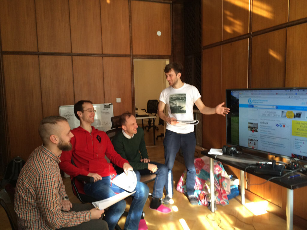 Three young men sitting in front of a screen, with a fourth standing and explaining something during an English class.